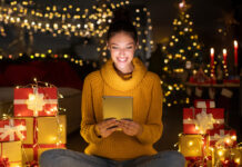 Best Tech Gifts in 2024 Smiling woman using digital tablet sitting in decorated room among gift boxes and garland, surfing internet and ordering presents