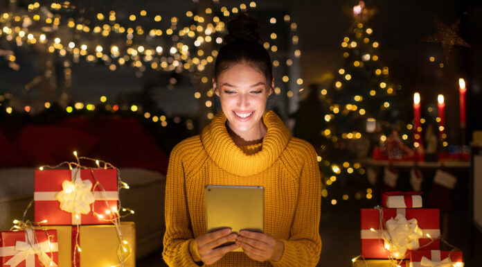 Best Tech Gifts in 2024 Smiling woman using digital tablet sitting in decorated room among gift boxes and garland, surfing internet and ordering presents